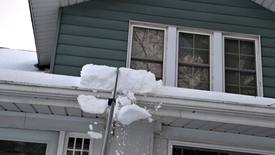 A silver roof rake removes snow from a faded teal 2-story house with a white roof