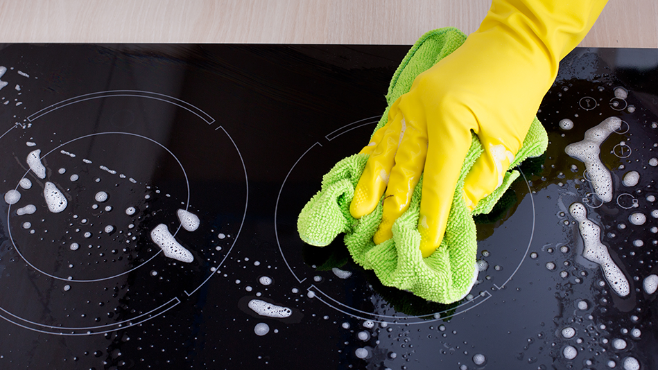 A hand in a yellow cleaning glove uses a neon green cloth to clean a soapy cooktop