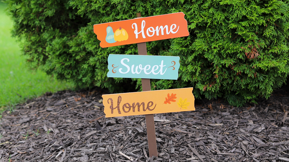 A fall-themed "Home Sweet Home" yard sign sticks out of mulch in front of a shrub