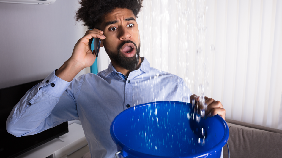 A black male shares surprise through the phone as water pours into a bucket from the ceiling above