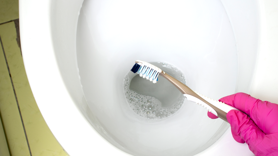 A pink-gloved hand holds a toothbrush over a now-clean toilet bowl that has bubbles in the water at the bottom of the bowl after cleaning; the floor tiles are a faded lime green