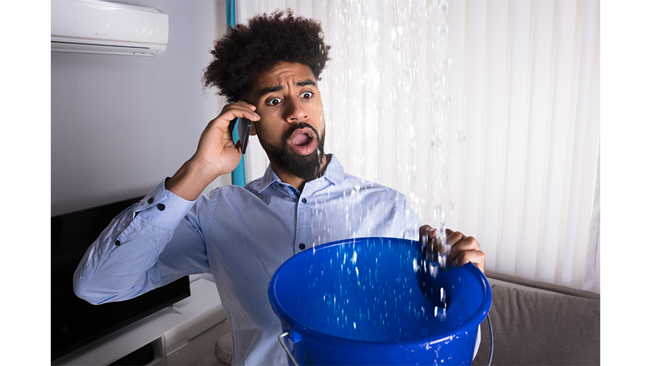 A black male holds a bucket to catch water coming from his ceiling while calling a plumber