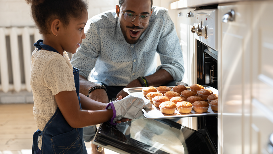 A Black man looks wowed as his young daughter pulls a tray of muffins out of the oven 