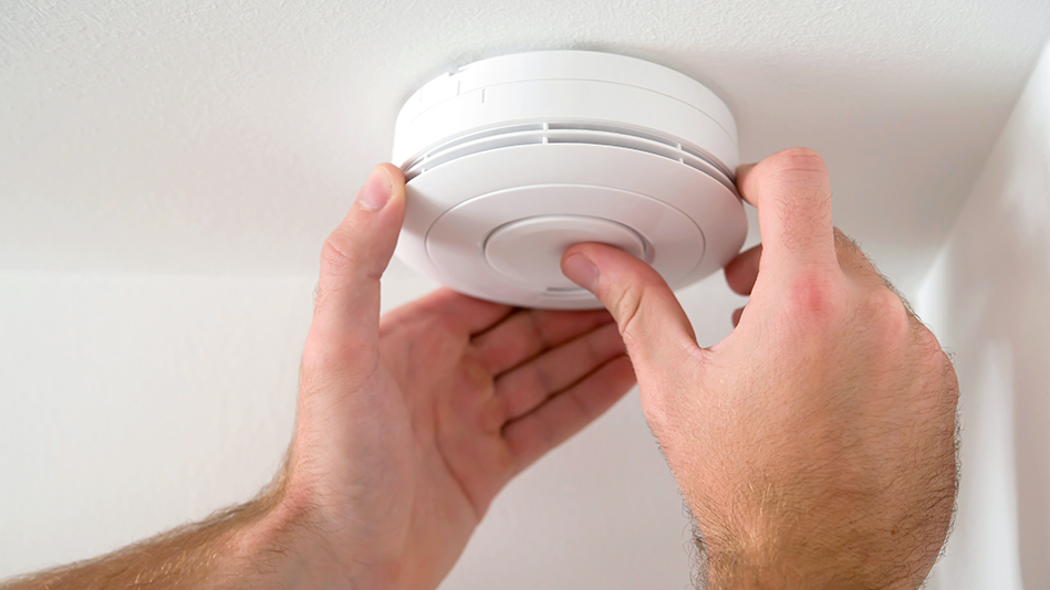 A white man's hands hold and press a smoke detector on a white ceiling next to a white wall