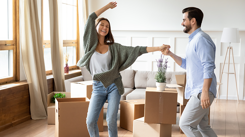 A Hispanic woman in a green sweater, white camisole, and light blue jeans is twirled by a Hispanic man in glasses, a blue button-down shirt, and gray slacks in their monochromatic tan living room with 6 large cardboard boxes and 2 plants behind them