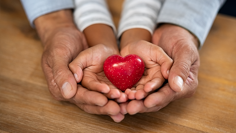 A red ceramic heart with small white dots sits in the hands of a Blended child in a gray-and-white long-sleeved shirt inside the hands of a Blended grandfather in a gray button-down shirt (forearms only) on a light wood table