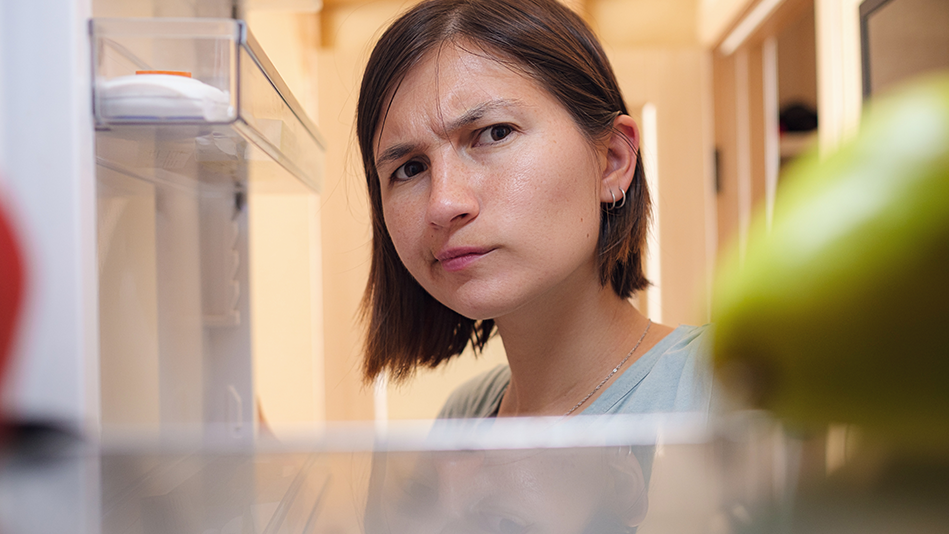 Blended woman with a brown bob haircut wearing a blue-gray shirt, 2 small silver earrings in her left earlobe, and a dainty silver necklace stares quizzically at the shelf of a fridge, which contains a pear on the right side and a red piece of produce on the left 