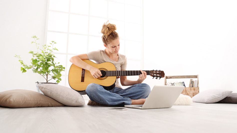 White woman playing guitar on the floor surrounded by neutral-colored pillows, a plant, and a toolbox