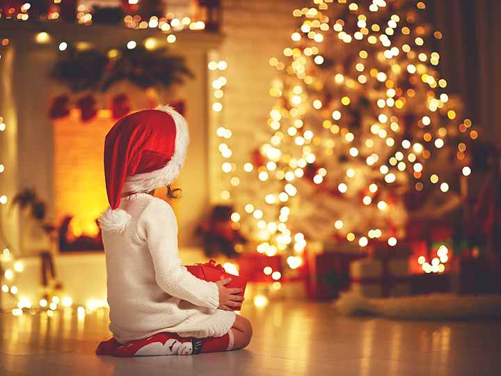 A small girl in the foreground wearing a white sweater dress, a red and white Santa hat, and red long socks with white snowmen on them sits on her knees on a wooden floor as she gazes at the background at an illuminated white fireplace adorned with 5 red stockings and greenery and a small burgundy and white doll that sits near the hearth to the left of a lit white Christmas tree with red, white, and green presents wrapped below it resting on a furry white rug  