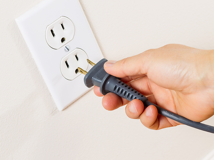 A light-skinned right hand reaches to plug a gray 120 V electrical cord with bronze prongs into a white vertical outlet  
