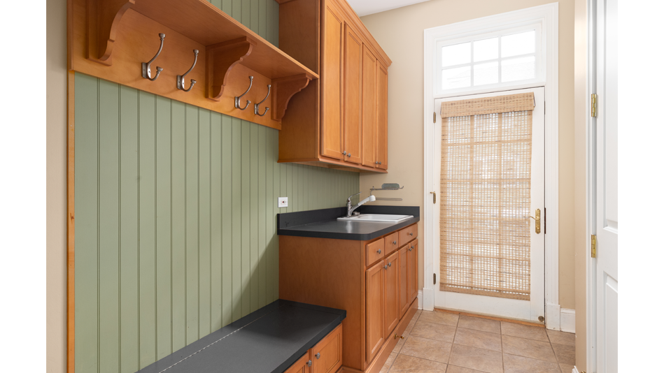 A mudroom with one tan wall and medium-toned wood cabinets and hook board mounted in front of olive green wood paneling. Between the two rows of cabinets sits a white-bowled sink with a black countertop.