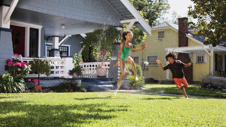 A little Black girl and a little Black boy frolic through the sprinkler on their front lawn; their lawn sits in front of a big, light blue house with a white veranda, trim, and accents; the veranda carries beautiful green and pink plants, with pink and green foliage in front; the neighbors' house is of similar size and shape, and it is painted a pale yellow; there is a big tree that casts a lovely shadow between the two houses