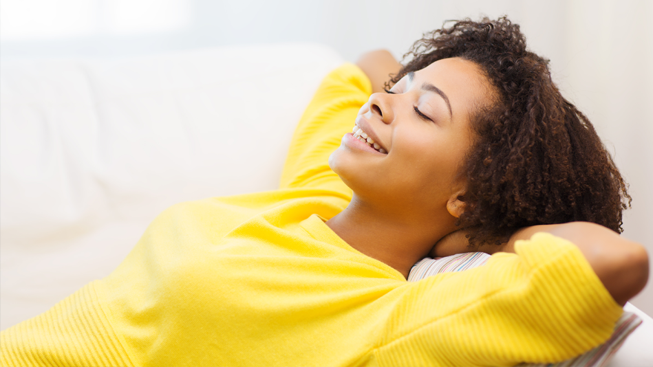 A Black woman with natural hair relaxes joyfully in a bright yellow half-sleeved sweater with horizontal ribbing on the sleeves and torso; she rests on a multicolored, vertically pastel-striped pillow on an all-white couch