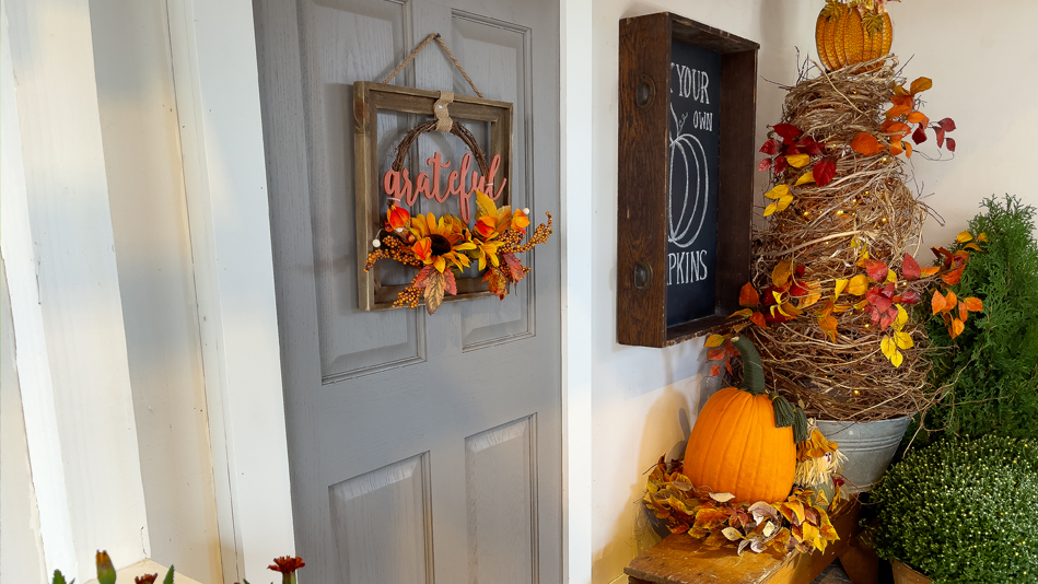 A white front porch with a gray front door is decorated for fall: From the door hangs a framed floral wreath that says, "grateful"; to the right of the door is a chalkboard sign reading "Pick Your Own Pumpkins" inside a vertical wooden box hung on the wall; below it is a bench that holds a pumpkin nestled in a wreath of leaves to the left of a pumpkin-shaped piece of décor resting on a tall spiraled nest of straw and leaves; in front of the bench sit two shrubs; the top blooms of a small houseplant poke into the bottom left corner of the frame