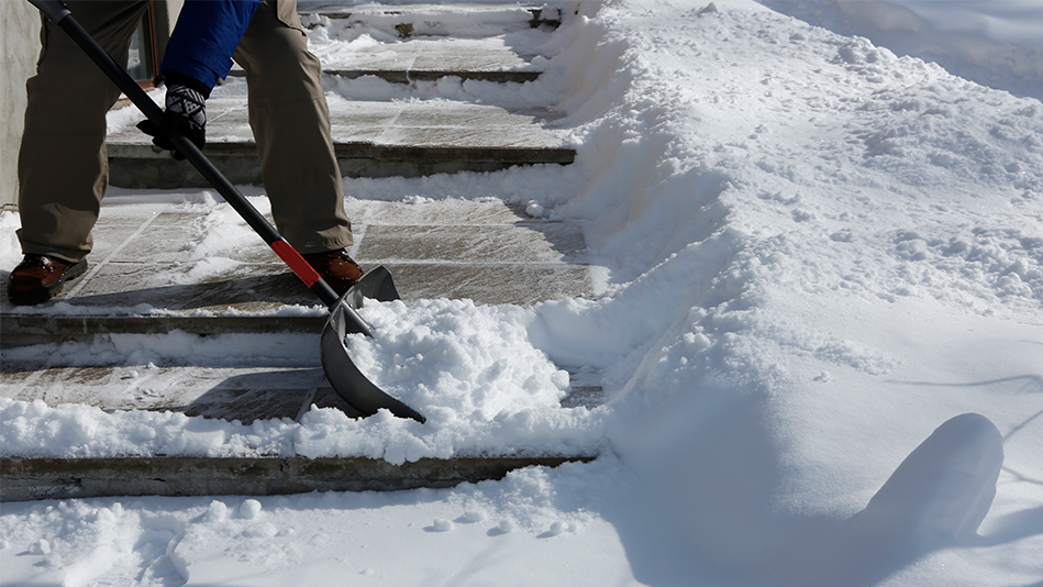 A person in gray-brown snowpants, a blue jacket, reddish-brown shoes, and black gloves uses a gray shovel with a black and red handle to clear a sidewalk of snow