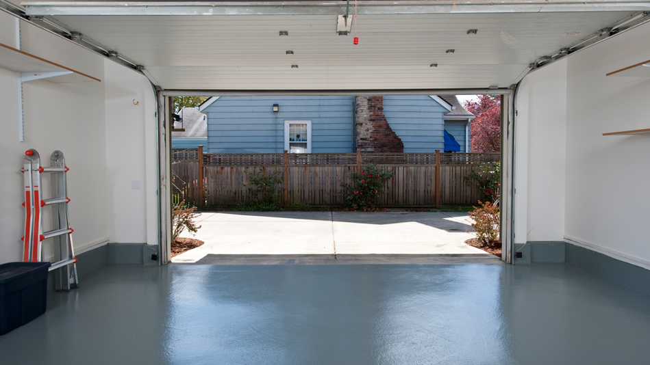 A clean and nearly bare garage with freshly polished and painted doors is open, showing the viewer the paved driveway in front of the 3 plants in view lining this side of the property; there is a light blue house with a brick chimney on the other side of the medium height fence