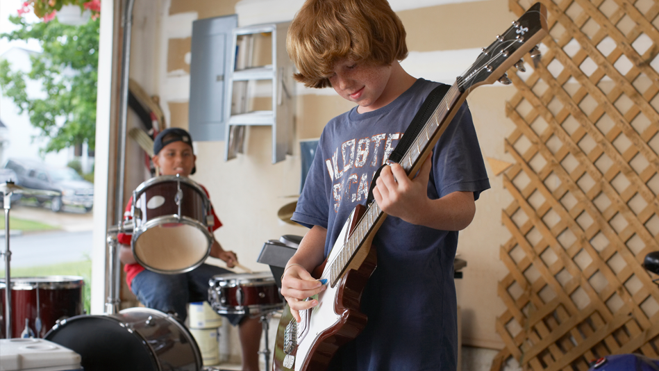 Two early adolescent males play instruments in a garage whose door is open, and whose walls display: A circuit breaker with a short metal stepladder hanging next to it on the left side of the wall, and a light wood trellis on the right side of the wall; the white early teenager is playing guitar, and the Hispanic early teenager is playing the drums  