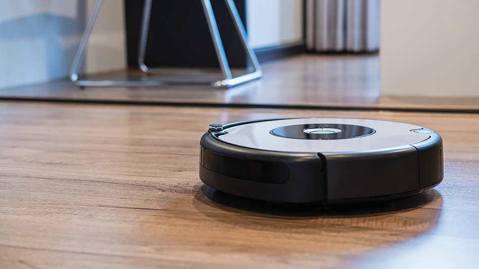 A black and silver robot vacuum cleans a hardwood floor with a metal chair in the background 
