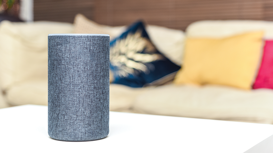 A gray cylindrical voice assistant device rests on a white coffee table in front of a tan couch adorned with 3 pillows: one navy with gold embroidery, one red, and one yellow 