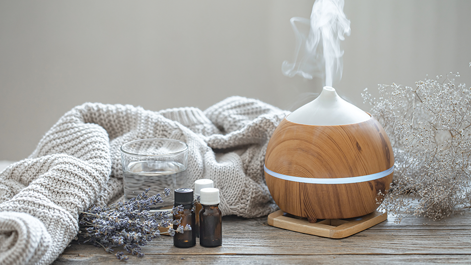 A spherical essential oil diffuser made of tan wood and white plastic rests on a wooden trivet and sits to the left of a bundle of baby's breath and to the right of an unfolded beige sweater, a clear, full glass of water, dried lavender, and small, brown bottles of essential oil 