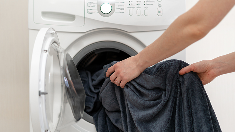 A white person with a small silver band on their left ring finger pulls a grayish-navy fleece throw blanket from a white washer
