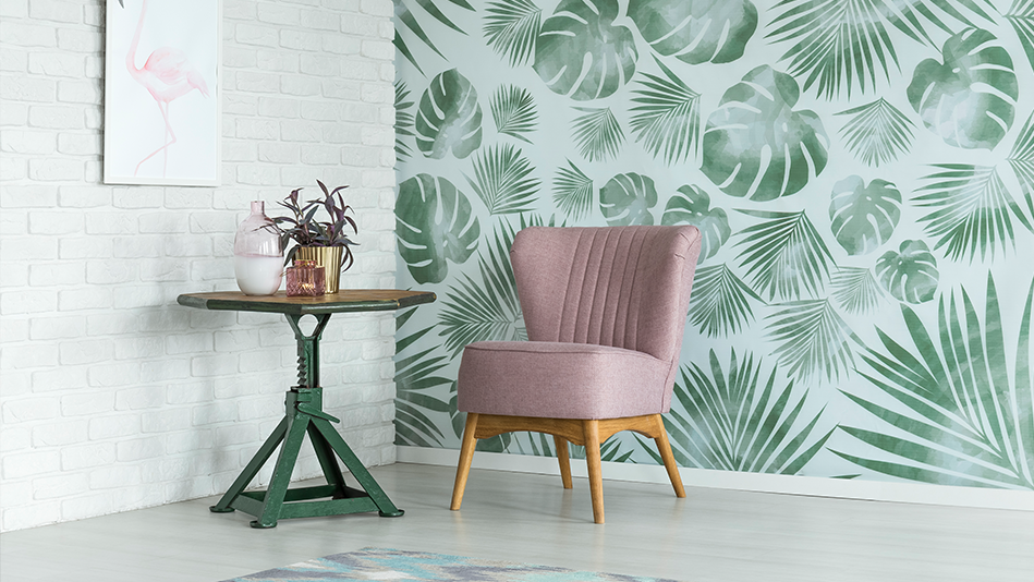 A blush-colored chair sits in a corner next to a green round coffee table; one wall is white brick with a flamingo picture, and the other wall is a green tropical leaf mural on a light blue background