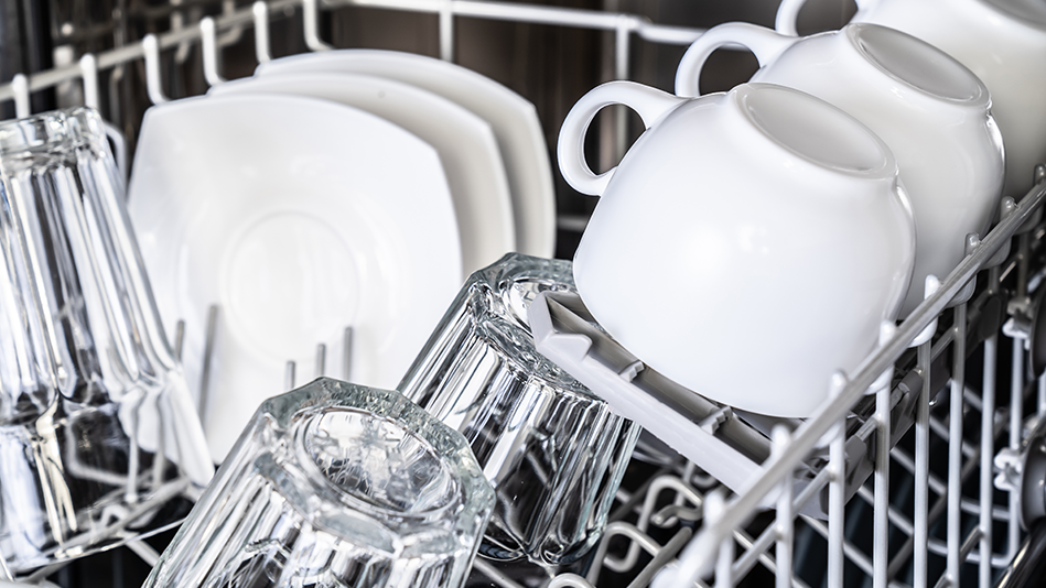 A top rack of a dishwasher is filled with clear drinking glasses, white upside-down cups, and white plates