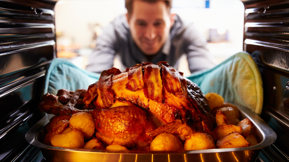 A white man reaches into an oven while wearing potholders to remove a bacon-wrapped turkey that sits on a bed of roughly chopped potatoes
