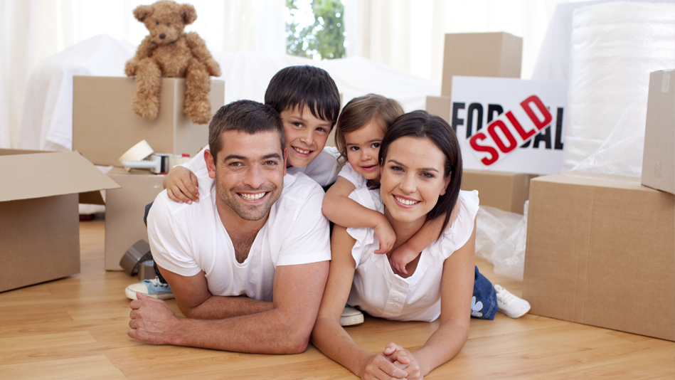 A family smiles together on the floor, surrounded by moving boxes and a SOLD sign
