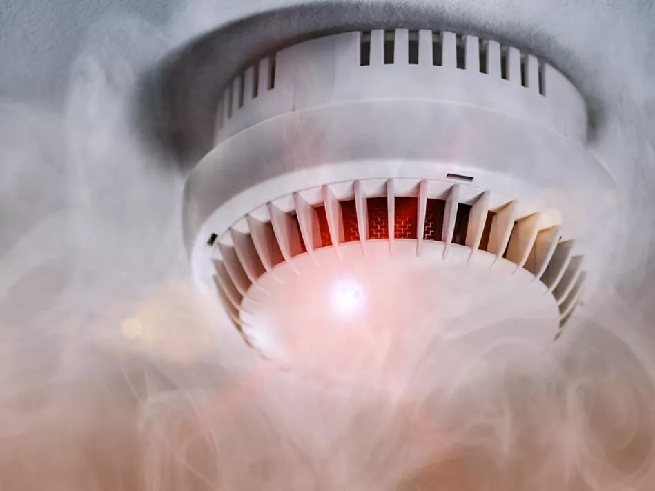 A white smoke alarm with a red light on and smoke around it