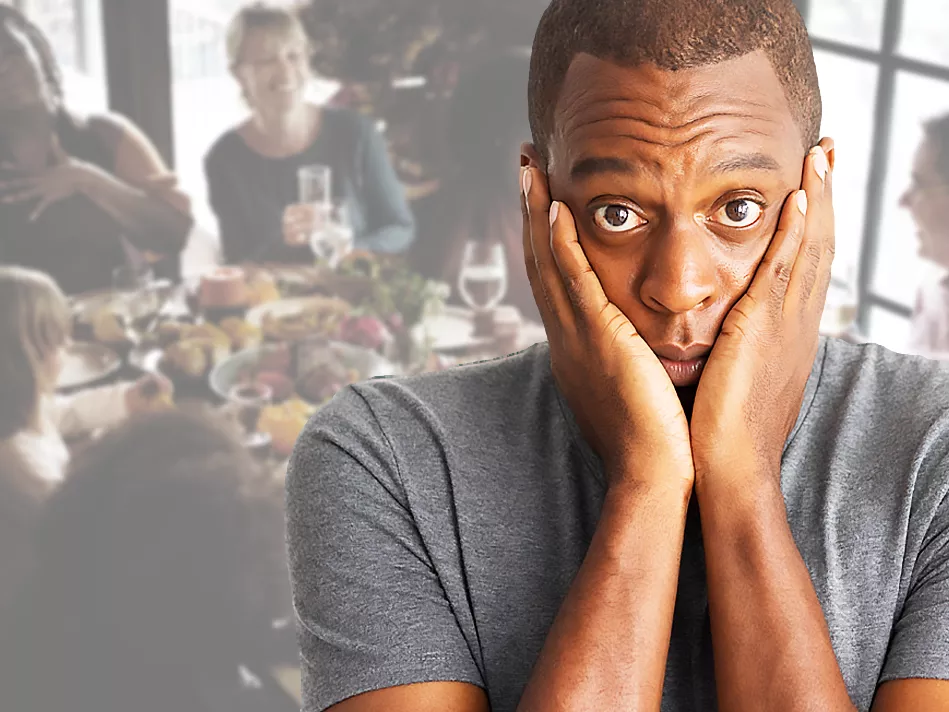 Black man with hands in a "V" over his face with people gathered at a dinner table in the background