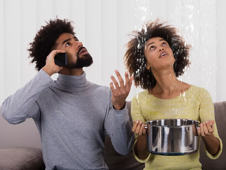Black man and woman on a couch looking up at the ceiling, with the man on a black phone and the woman holding a metal pot to catch the water falling directly above her 