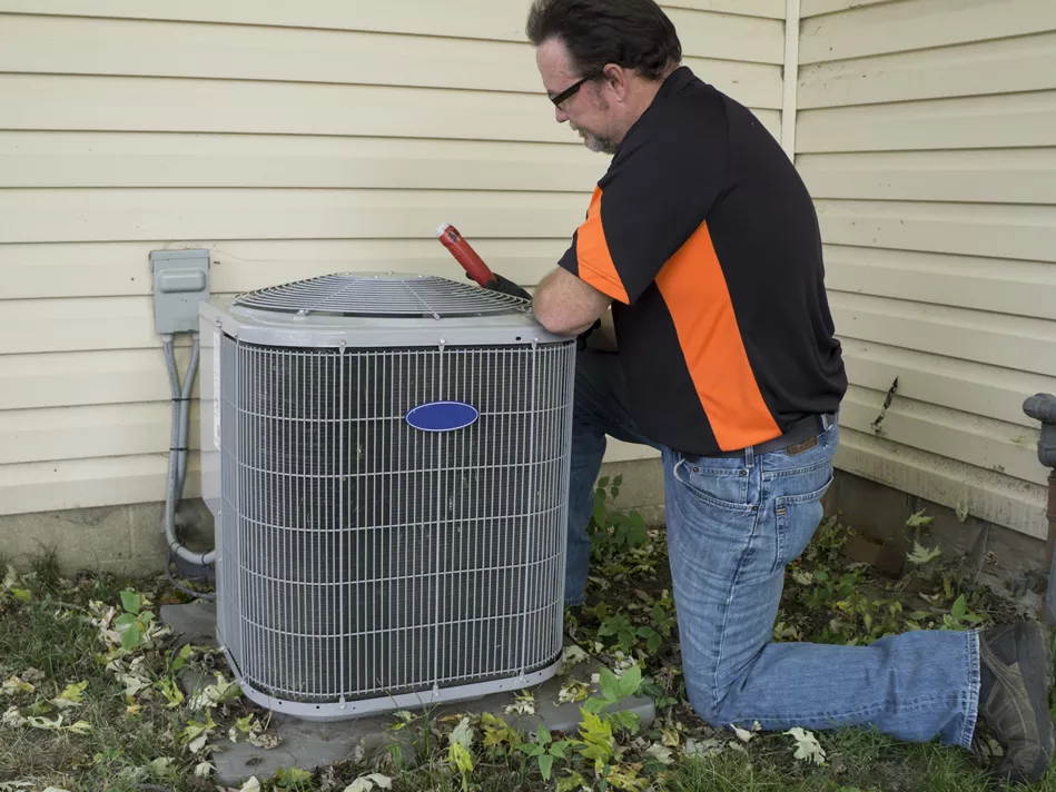 White man in glasses and a black-and-orange shirt and jeans kneels on the side of an air conditioner unit