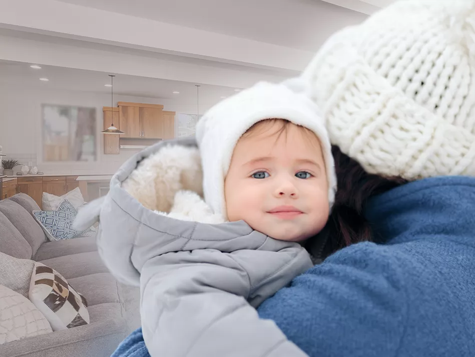 Woman in a blue coat and white hat holds a toddle in a gray coat and white hat in front of an open kitchen and living room 