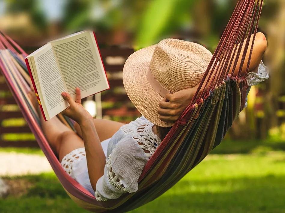 Blended woman in a tan beach hat and white sundress reading a book in a red hammock