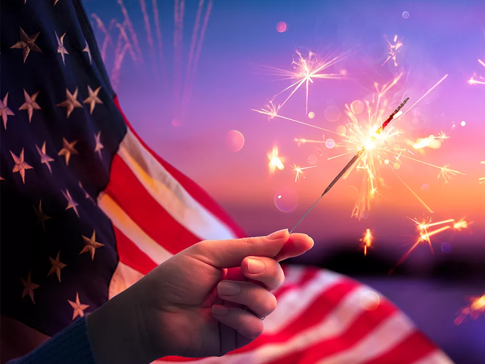 A hand waving a lit sparkler in front of an American flag at sunset