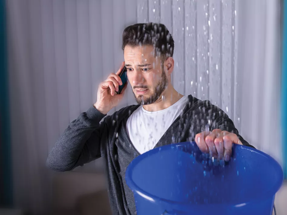 Hispanic man on the phone holding a blue bucket to catch water from a leak