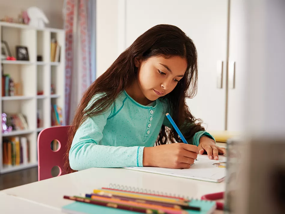 Hispanic girl in a green shirt writing on a sheet of paper at her desk with a full white bookcase in the background