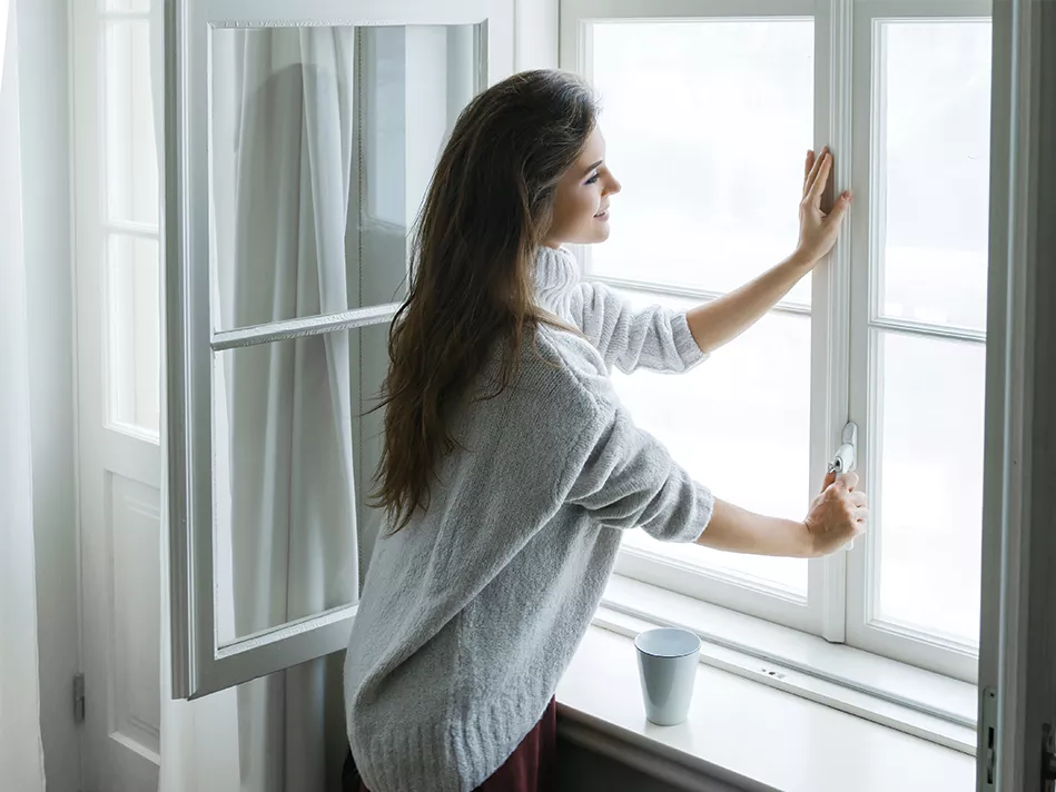 FOR WEB-White brunette woman in a sweater looking out a window with light curtains