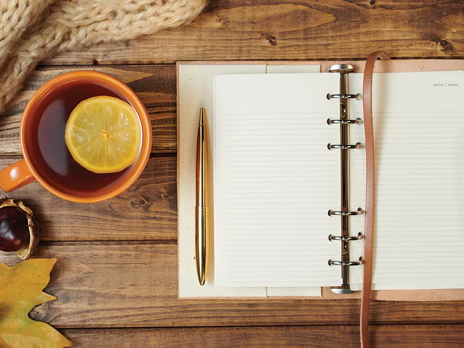 An orange cup with tea and a lemon slice in it next to an open blank journal with a pen on a wooden table