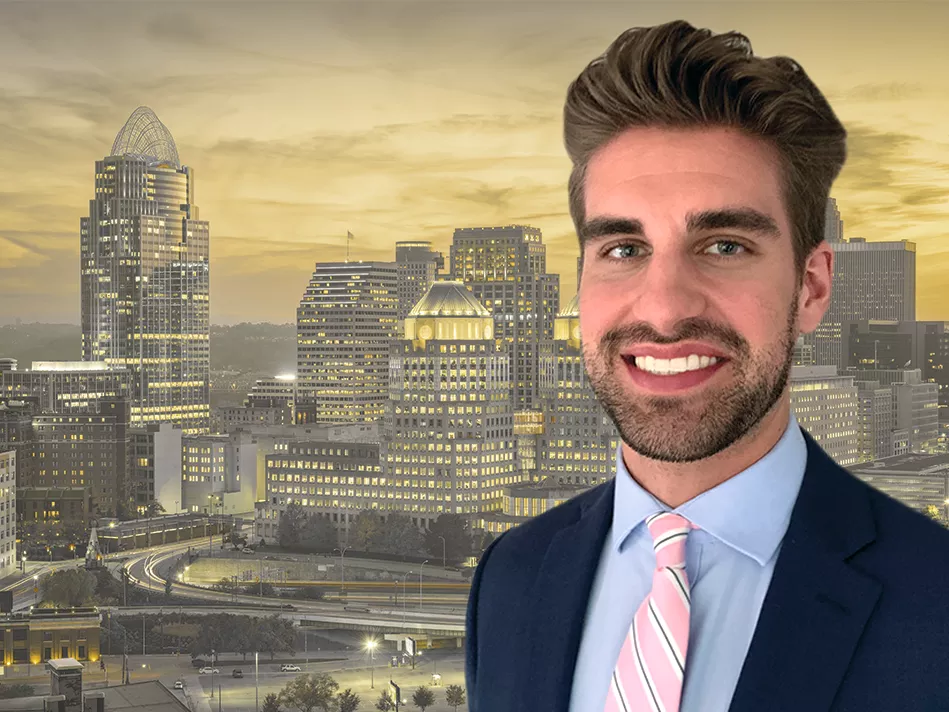 White adult male in a suit smiles in front of a cityscape