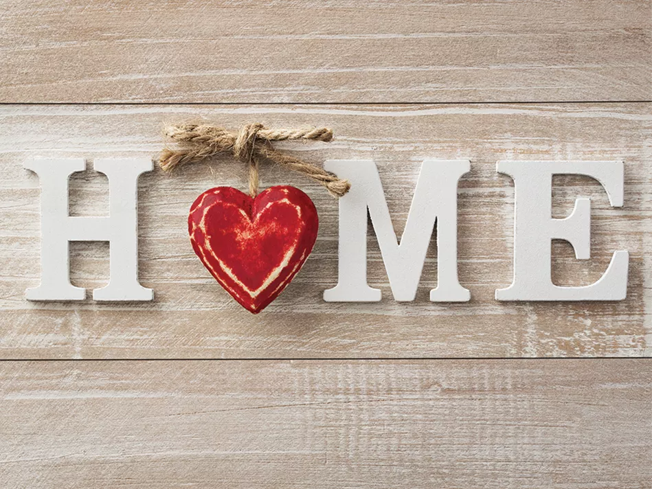 White capital wooden letters spell out "HOME" with a red wooden heart hanging from a bow-tied piece of twine standing in for the "O" on a tan-gray wooden table
