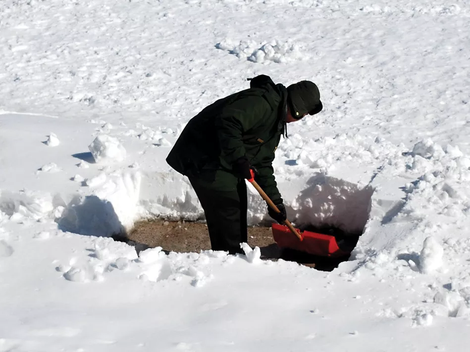 A person in a black hat, coat, pants, and gloves uses a red and wooden shovel to scoop foot-high snow around him 