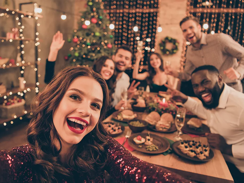 A brunette white woman wearing red lipstick and a sparkly dress takes a selfie of her and her friends, 2 white women, 2 white men, and 1 Black man, who are all dressed up at a long table full of assorted platters of food and a few flutes of wine; in the back left corner is a Christmas tree, and to the left of that is a wood and metal shelving unit decorated with LED holiday lights; the 2 windows at the back are covered in vertical string lights 