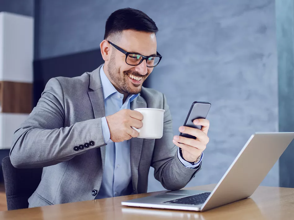 White male smiles over his coffee at his cellphone, in front of his laptop