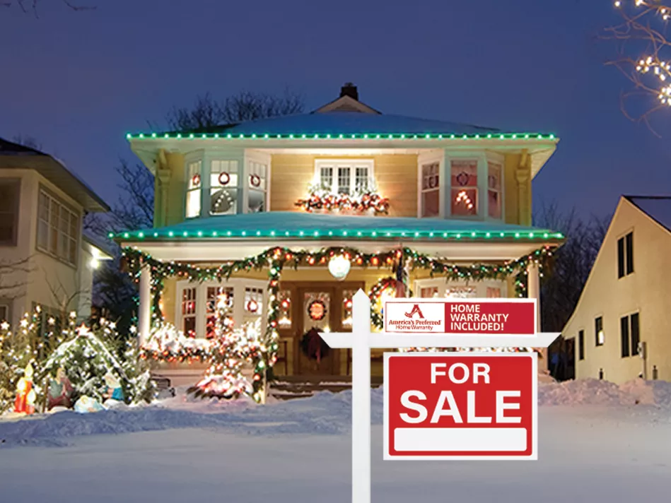 A two-story yellow home with white windows is fully decorated with green, red, and white Christmas lights, garlands, and a Nativity scene out front on the left; the front yard is covered with snow, and there is a red and white "For Sale" sign out front on a white post with a red and white rider sign that has the full APHW logo on the left side in red on a white background, and the words "HOME WARRANTY INCLUDED!" in white on a red background on the right side; it's flanked by 2 houses w/ trees behind 