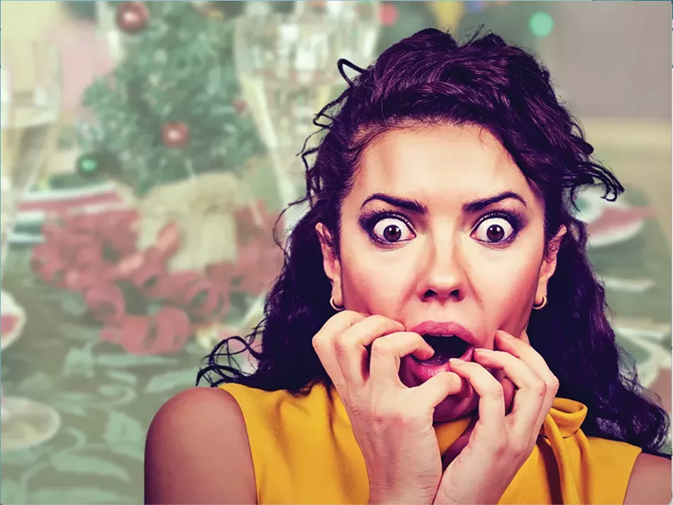 A curly-haired Blended woman in a yellow-gold top freaks out with her hands in her mouth in front of the background of a table covered in red and green Christmas decor 
