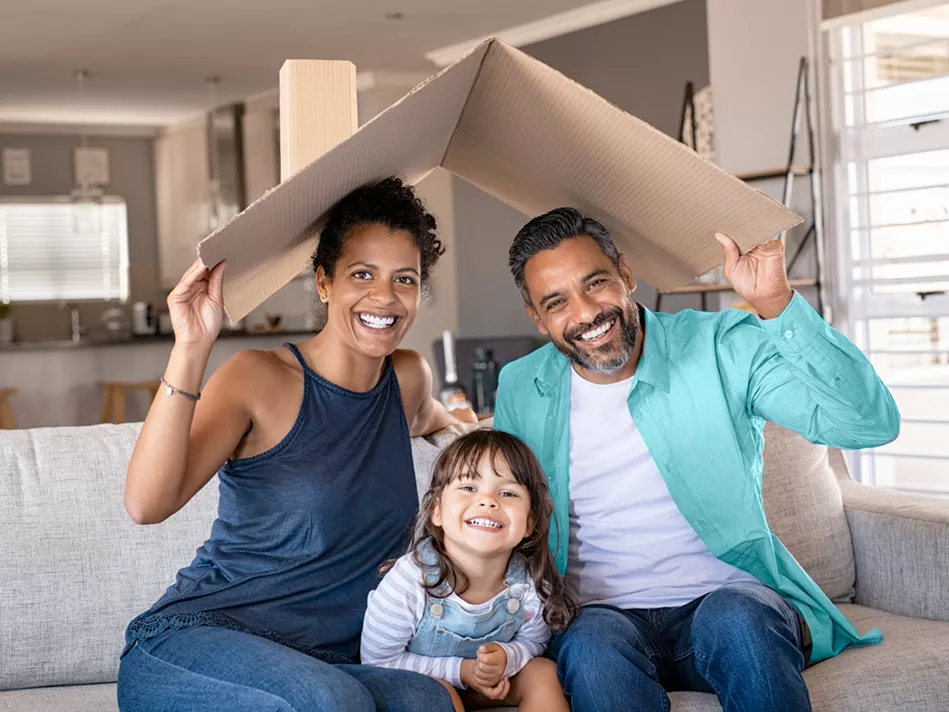 Blended man and woman hold up cardboard up as a "roof" over their heads with small Blended child sitting between them on a tan living room couch