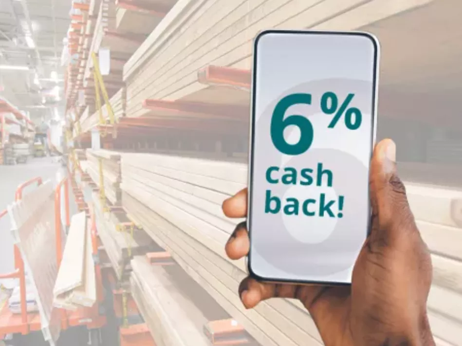 A Black right hand holds a smartphone whose screen bears the text "6% cash back!" in teal in over a white background with a large watermark of the number "6"; behind is the lumber aisle in a hardware store