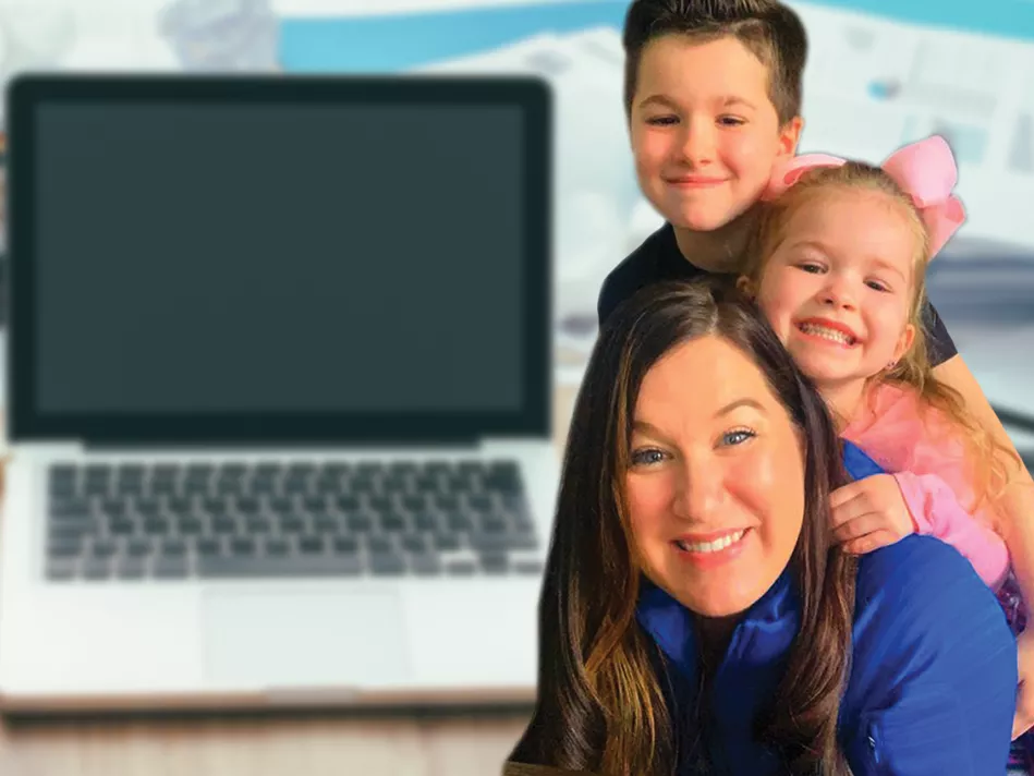 Fawna Farrell in a blue zip-up smiles with her daughter in a pink shirt and bow and her son in a black shirt smiling behind her; the background is an open silver computer on a light wood desk in front of a stack of papers with text and graphs